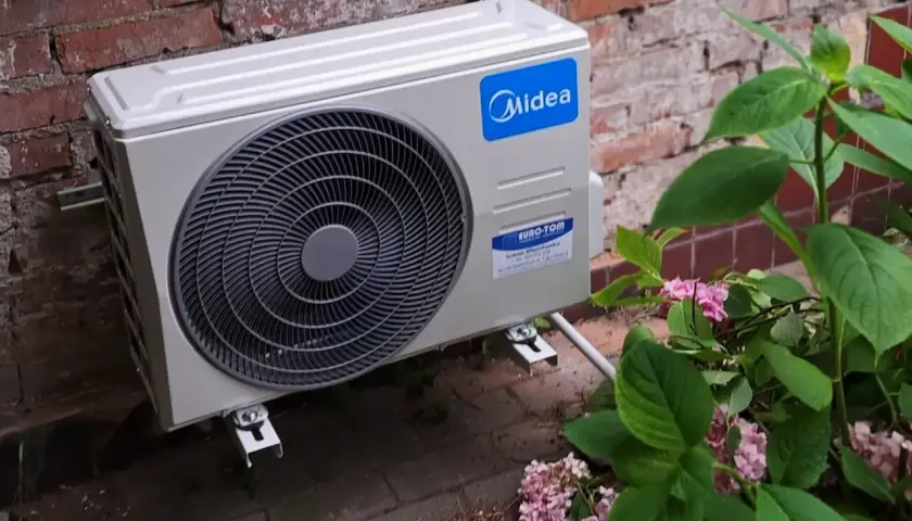 Jednostka zewnętrzna klimatyzatora Midea zamontowana na ceglanej ścianie, obok krzewu z różowymi kwiatami.